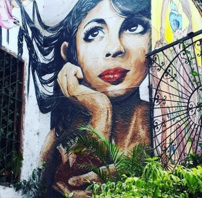 Street mural of a woman with flowing black hair and red lips, painted on a white building wall with tropical plants below.