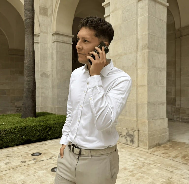 A young man in a white button-down shirt talks on a smartphone near arched stone architecture.