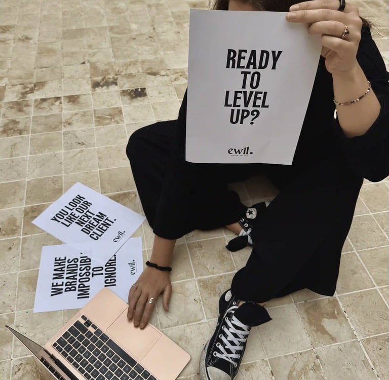 A creative professional holds a sign saying Ready To Level Up next to a laptop and marketing flyers.