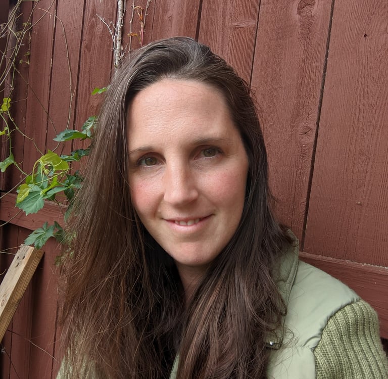 A smiling woman with long brown hair wearing a green vest against a rustic red wooden fence.