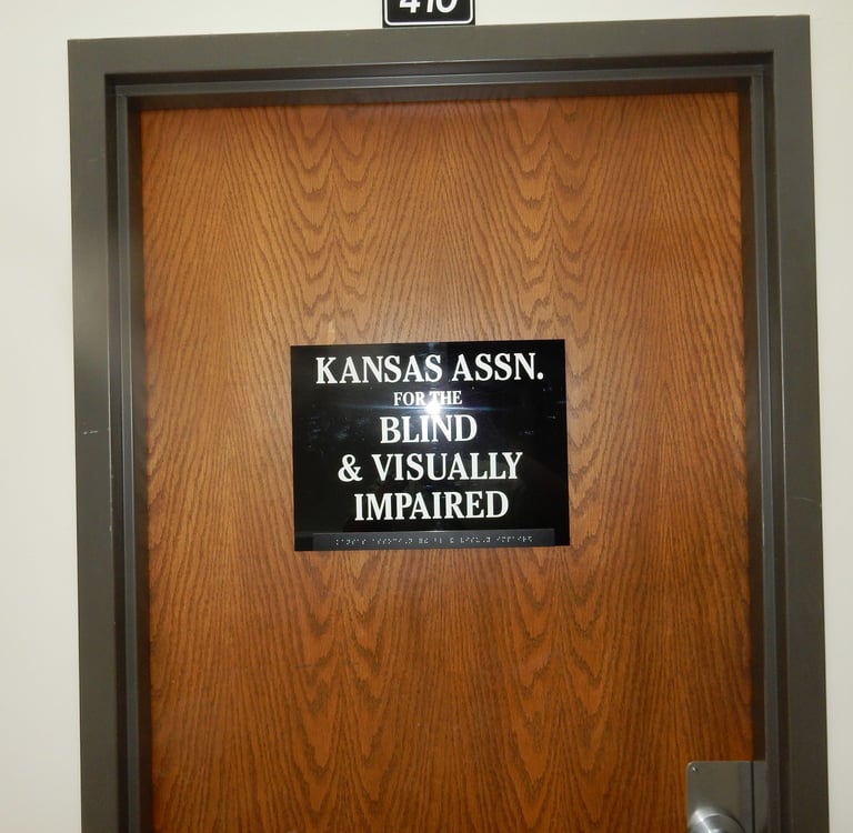 "A wooden office door with a black sign that reads 'KANSAS ASSN FOR THE BLIND & VISUALLY IMPAIRED