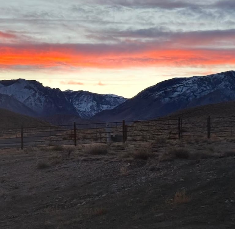 Vibrant orange sunset over snow-capped mountains behind a ranch fence in a high desert landscape.