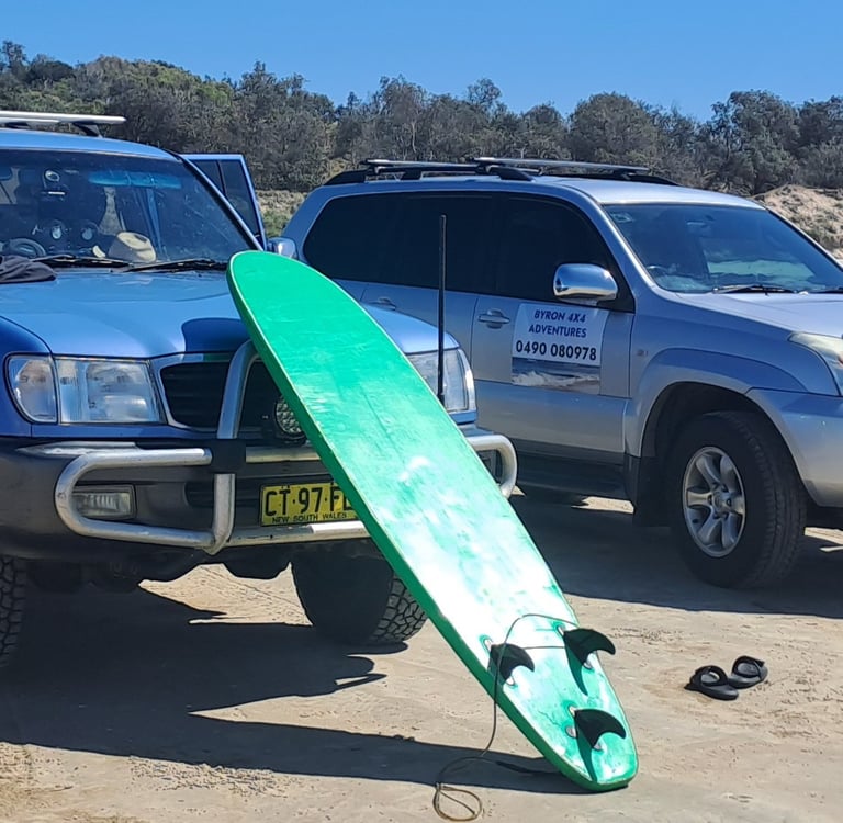 Two 4x4 off-road vehicles parked on a sandy beach with a green surfboard and camping chairs for Byron Bay adventures.