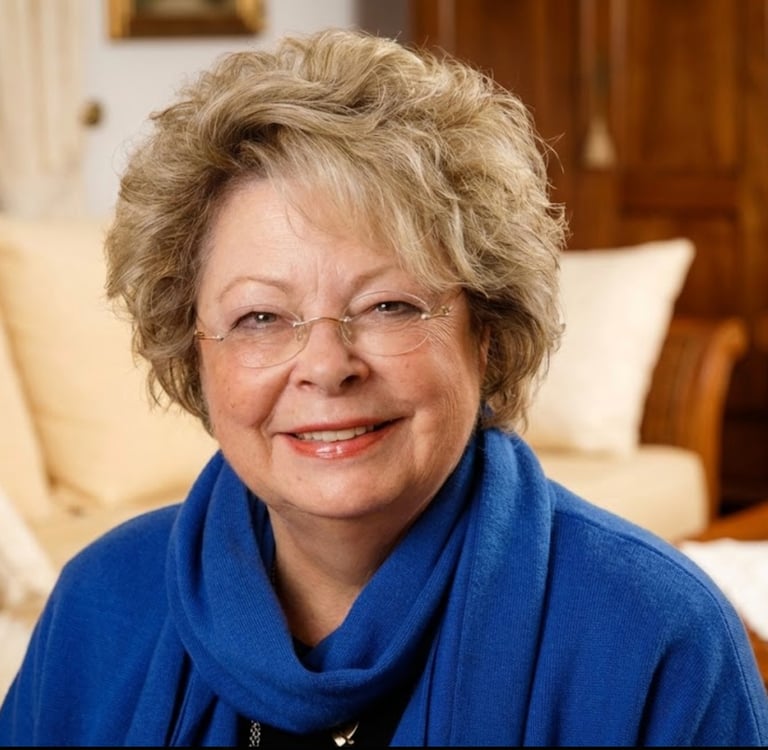 Smiling senior woman with short blonde hair and glasses wearing a blue scarf in a living room.