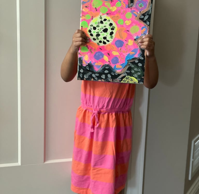 little girl with dark hair in a pink and orange dress holds colorful painting in front of her face