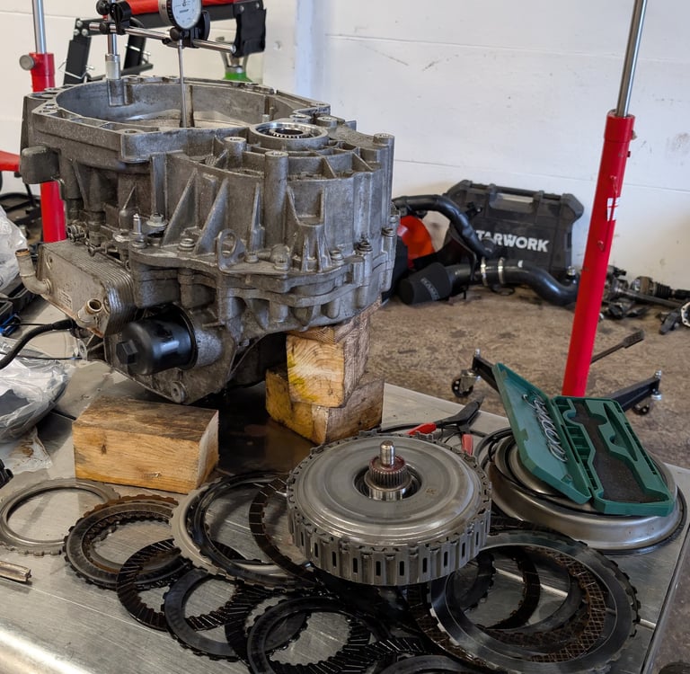 Automatic transmission rebuild with clutch plates and dial indicator tool on a metal workbench.