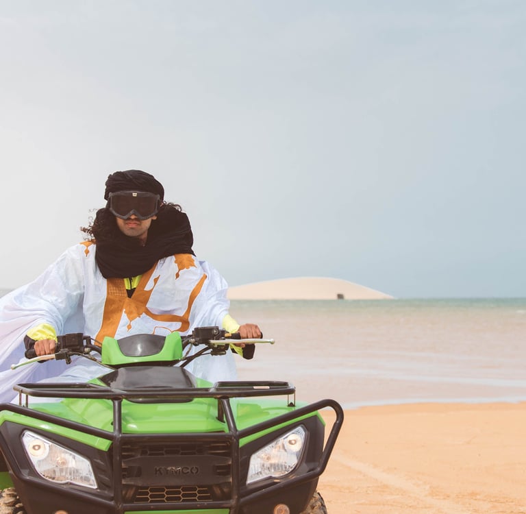 Man in traditional Sahraoui attire riding a lime green quad bike on the beach in Dakhla with the white dune in the background