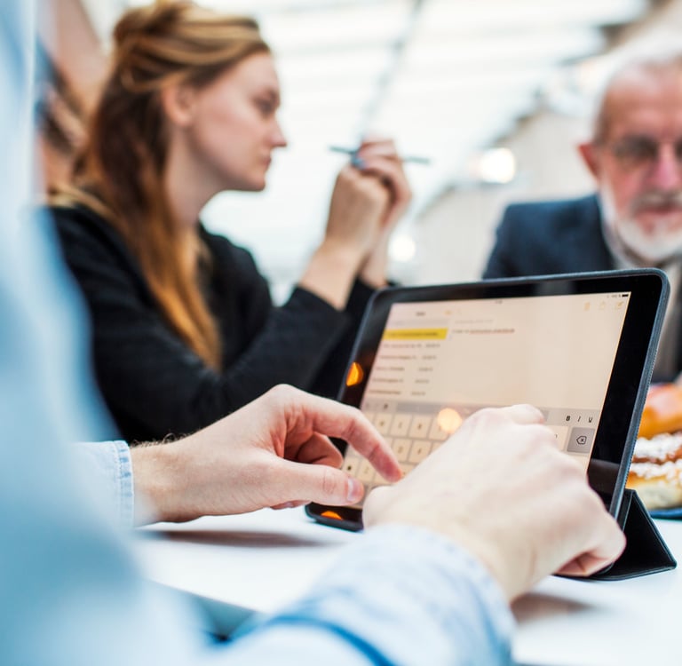 Person using a tablet during a meeting, with colleagues in discussion in the background.