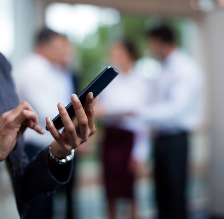 Person using a smartphone with colleagues conversing in the background.