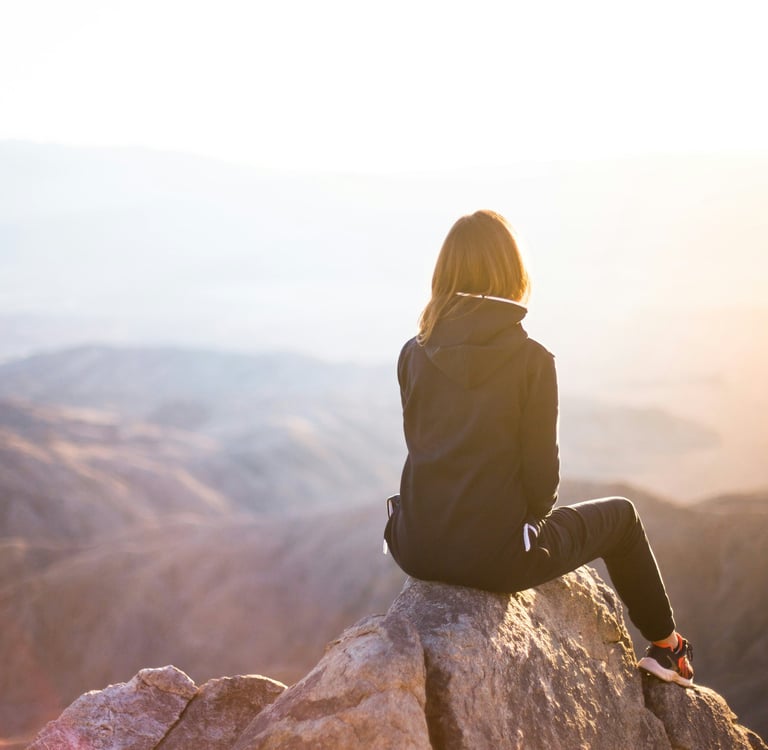 Woman sitting on mountain peak at sunrise, reflecting in solitude and enjoying peaceful nature view.
