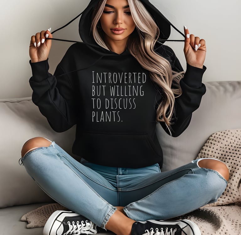 Woman in a black “Introverted but willing to discuss plants” hoodie sitting cross-legged indoors.