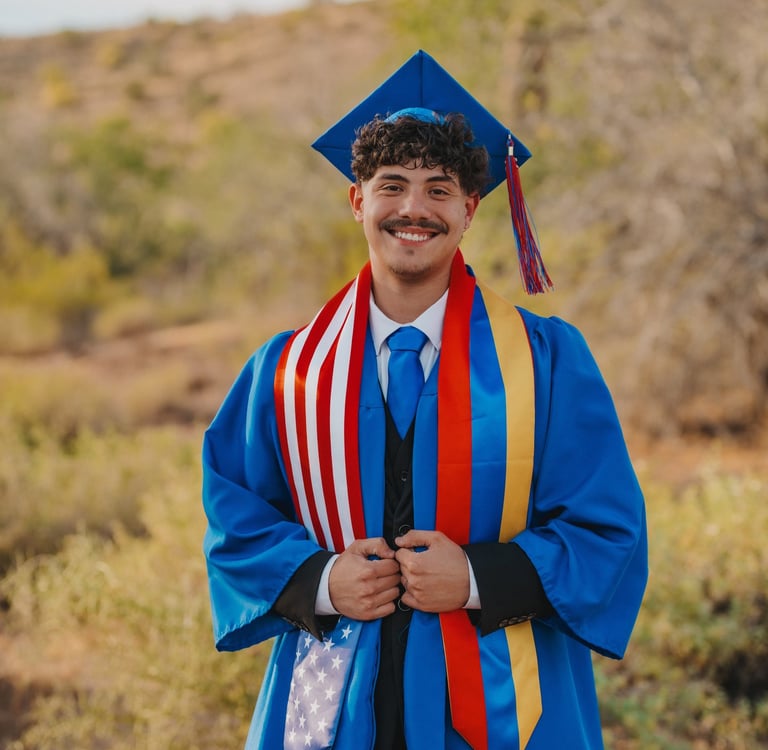 A smiling male graduate in a blue cap and gown wears a patriotic American flag stole for an outdoor portrait.