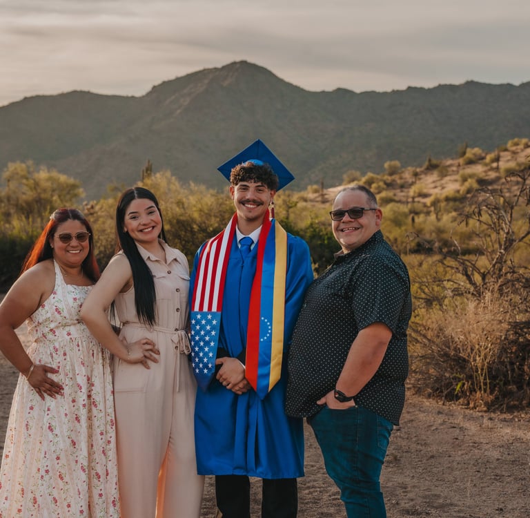 A graduate in a blue cap and gown stands with family in a desert landscape at sunset.