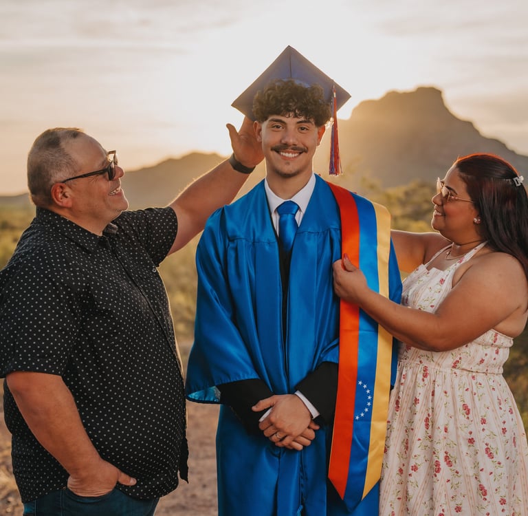 A graduate in a blue gown and cap stands with his parents at sunset in a desert landscape.
