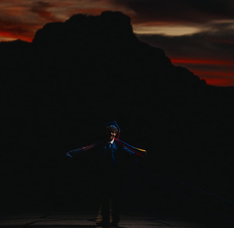 A graduate in cap and gown holds an American flag at sunset with mountain silhouettes.