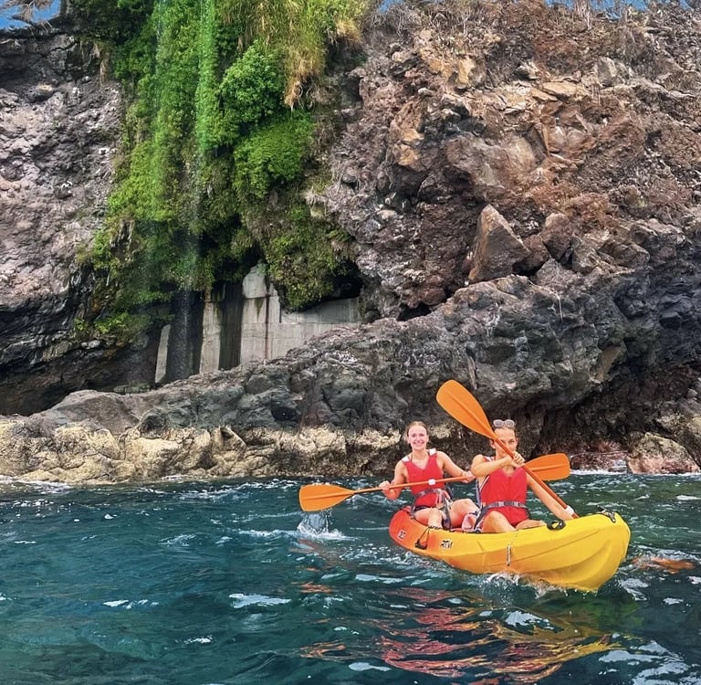 Two women in lifejackets paddling a yellow kayak past a green cliff waterfall on the Madeira coast