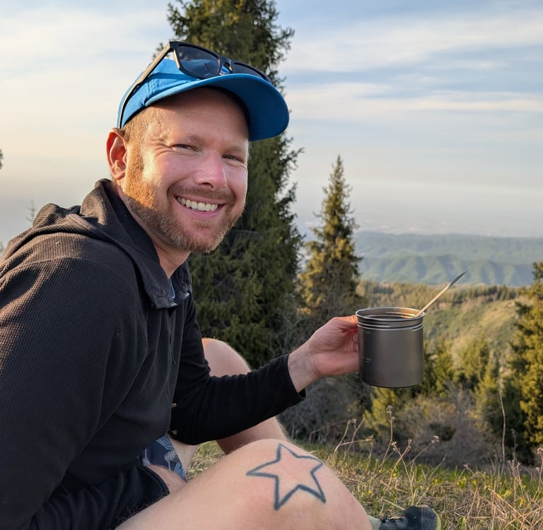 A smiling male hiker holding a Vargo BOT 700 mug while sitting on a scenic mountain ridge at sunset.