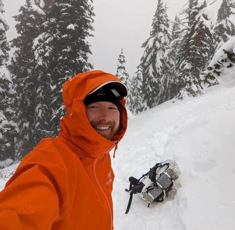A smiling man wearing an orange Arc'teryx jacket with a Hyperlite backpack lying in the snow.