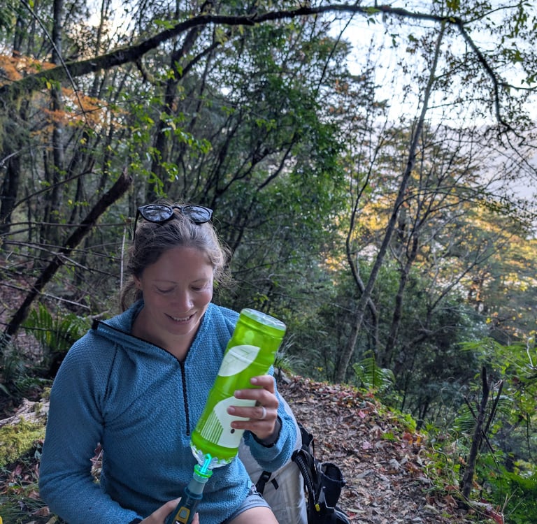 A female hiker filters water using a Katadyn Ultra UV filter into a green bottle on a forest trail.