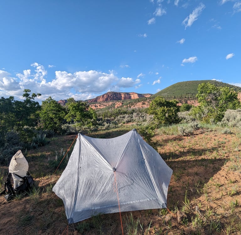 A backpacking tent pitched in a desert wilderness camp with red rock mountains under a blue sky.
