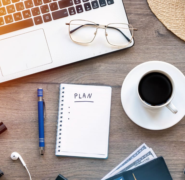 Uma caderneta de planos repousa numa mesa junto a notebook, lapiseira, xícara de café e óculos