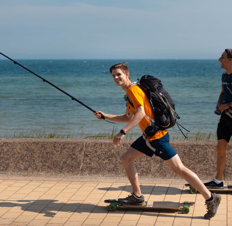 Zwei Männer fahren Longboard an der Küste mit Selfie-Stick. Lifestyle-Foto von Just Perspectives.