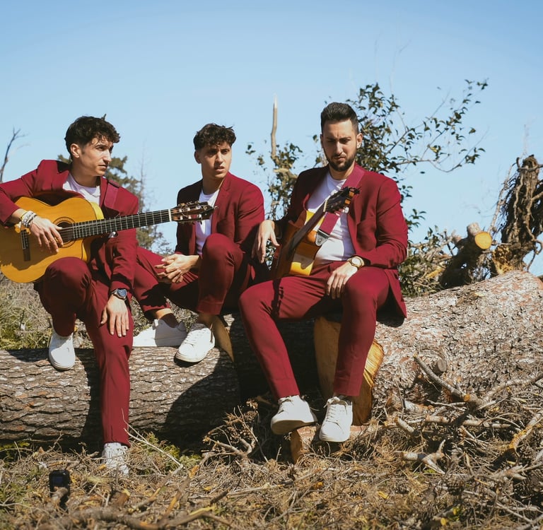 Three male musicians in burgundy suits playing acoustic guitars while sitting on a large log outdoors.