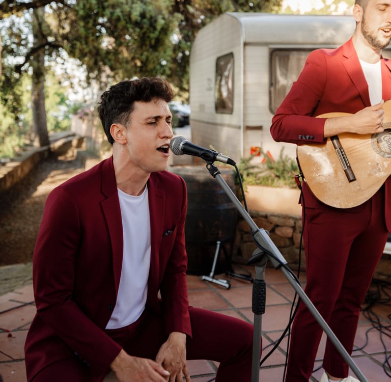 A live wedding band in burgundy suits performing outdoors with acoustic guitars and microphones.