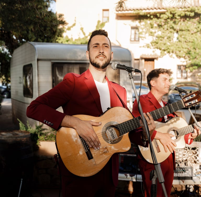 Live wedding musicians in red suits playing acoustic guitars and singing at an outdoor reception.