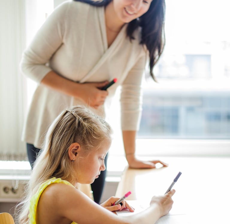 Mokytoja padeda vaikui. Mergaitė rašo į sąsiuvinį.