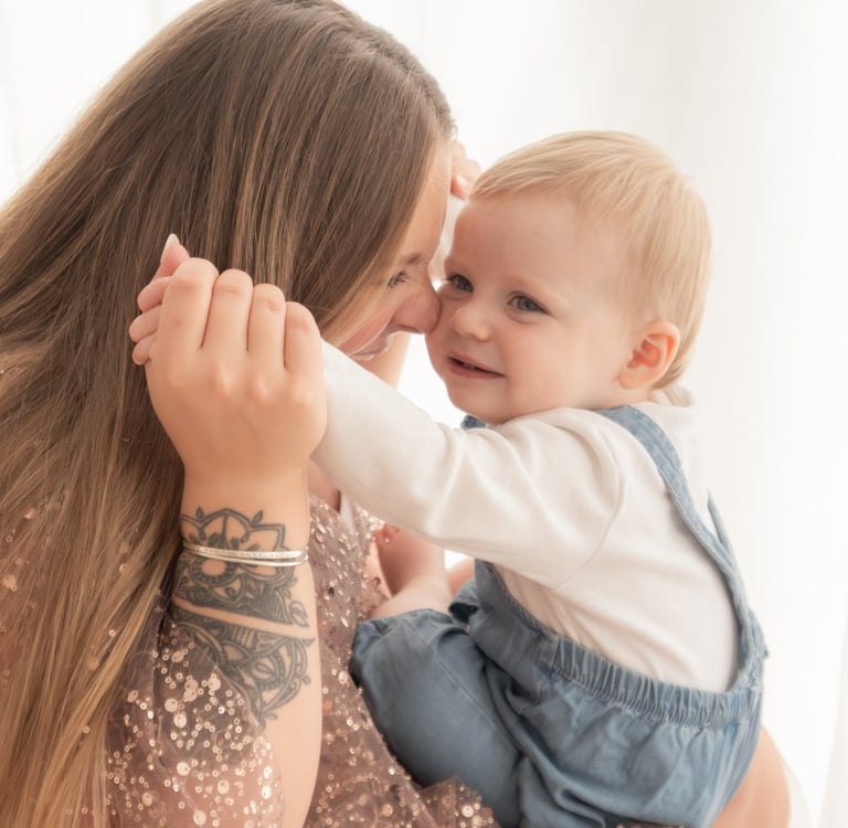 A smiling mother with a forearm tattoo holding her blonde toddler boy in blue overalls.