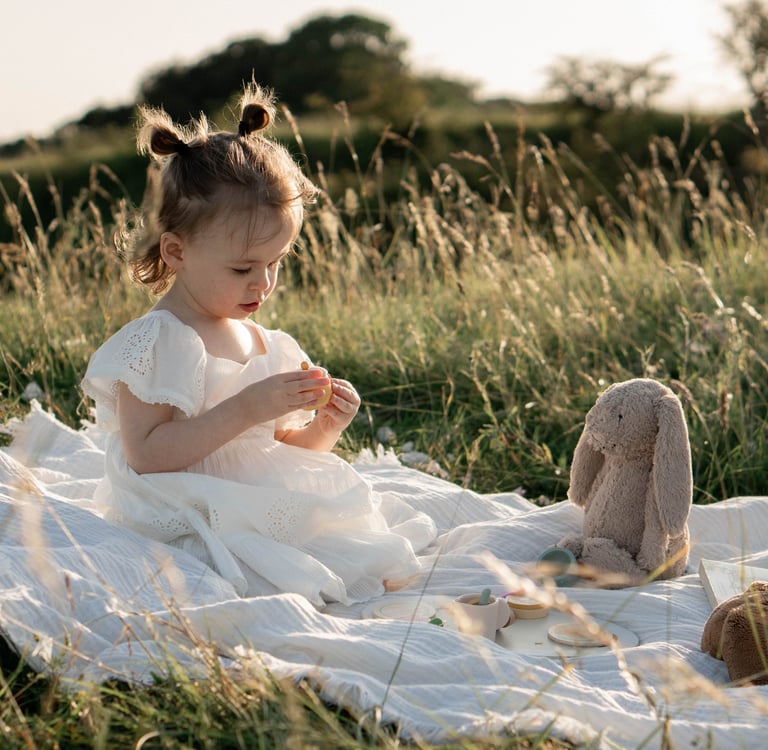 Toddler girl in white dress having a picnic with a stuffed bunny in a sunny grassy field.