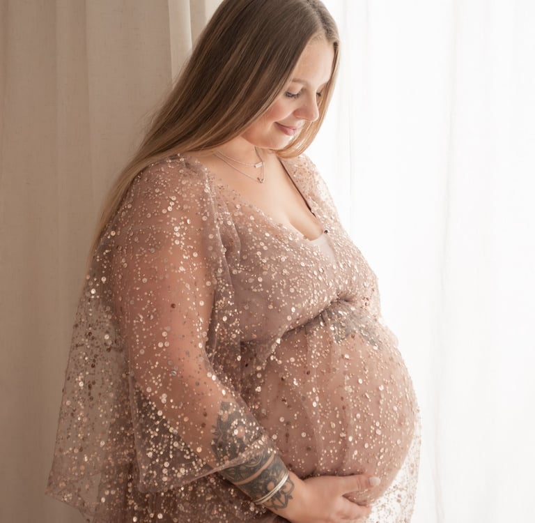 A smiling pregnant woman with tattoos wears a rose gold sequin maternity dress for a studio photo shoot.