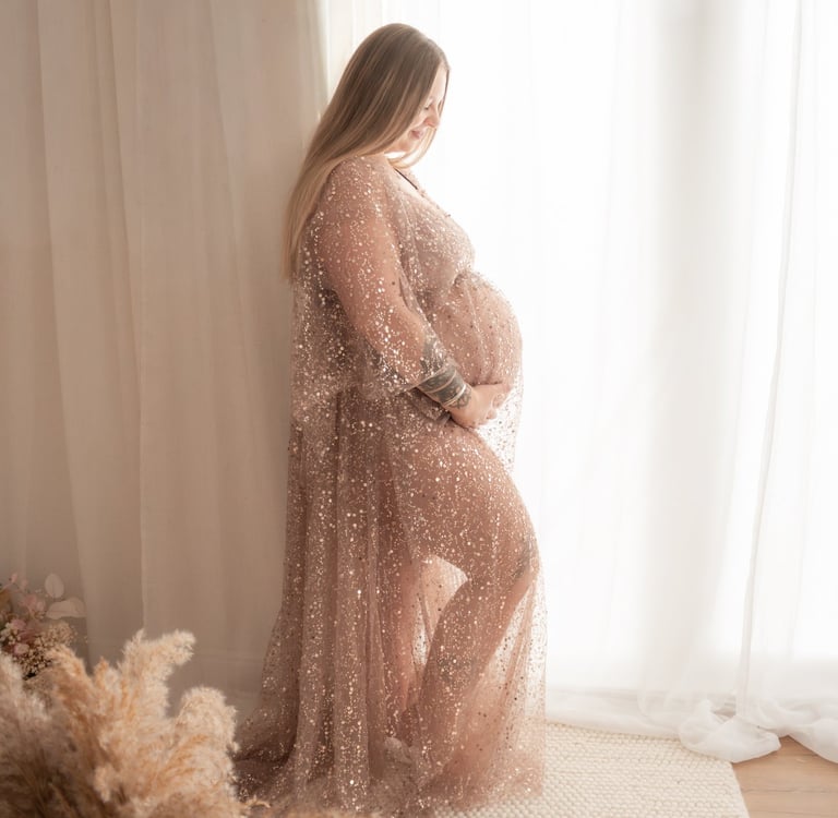 Pregnant woman in a sparkly maternity gown posing for a photoshoot by a window.
