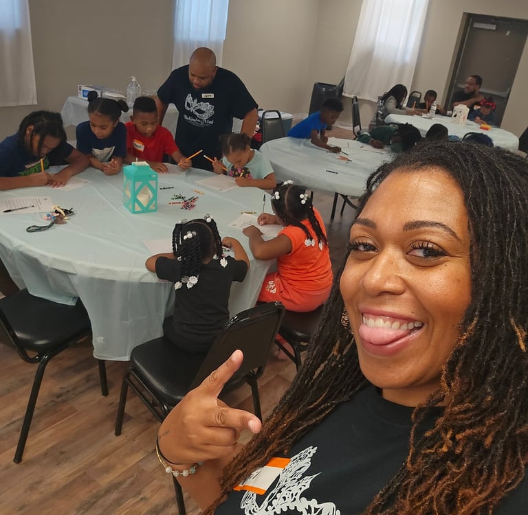 Smiling teacher takes a selfie while students engage in a creative art activity at a youth community center.