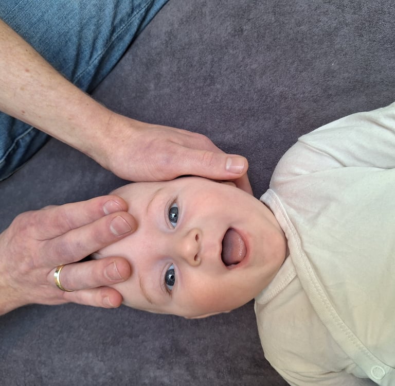 een baby ligt op zijn rug en lacht terwijl 2handen zijn hoofd vasthouden.