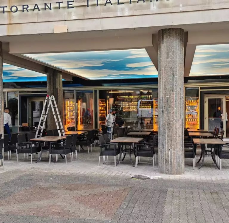 Outdoor patio of an Italian restaurant with modern chairs, stone pillars, and a cloud-painted ceiling.