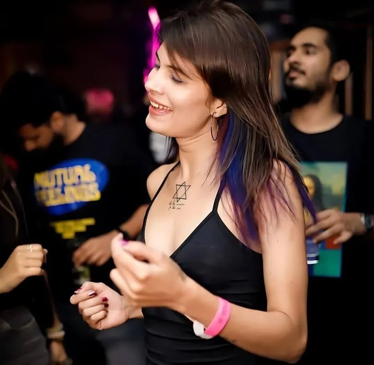 Smiling woman with purple hair highlights and a chest tattoo dancing at a crowded night club party.