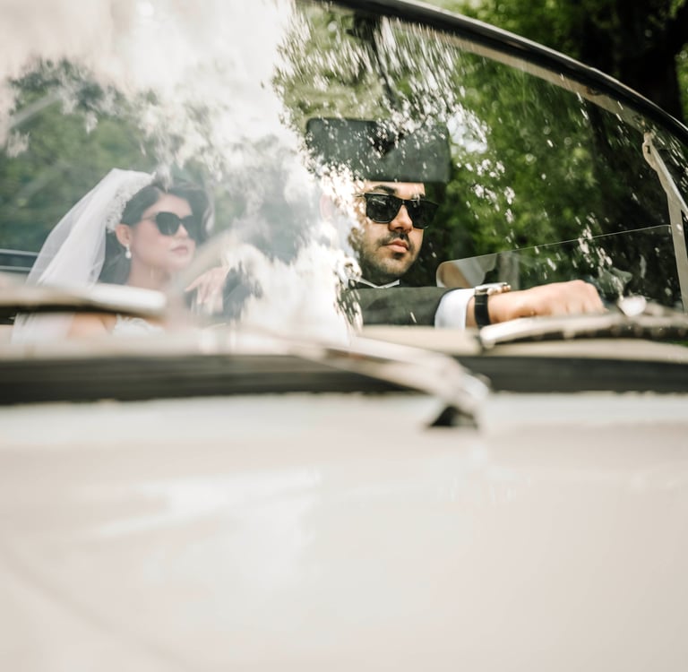 Bride and groom wearing sunglasses driving a classic car, captured by Fred Art Studio.