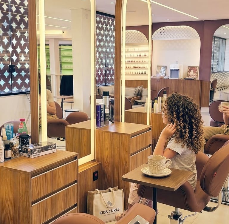 Menina cacheada admirando o resultado do seu tratamento no espelho, com a mãe sorrindo ao fundo no We Hall Beauty.