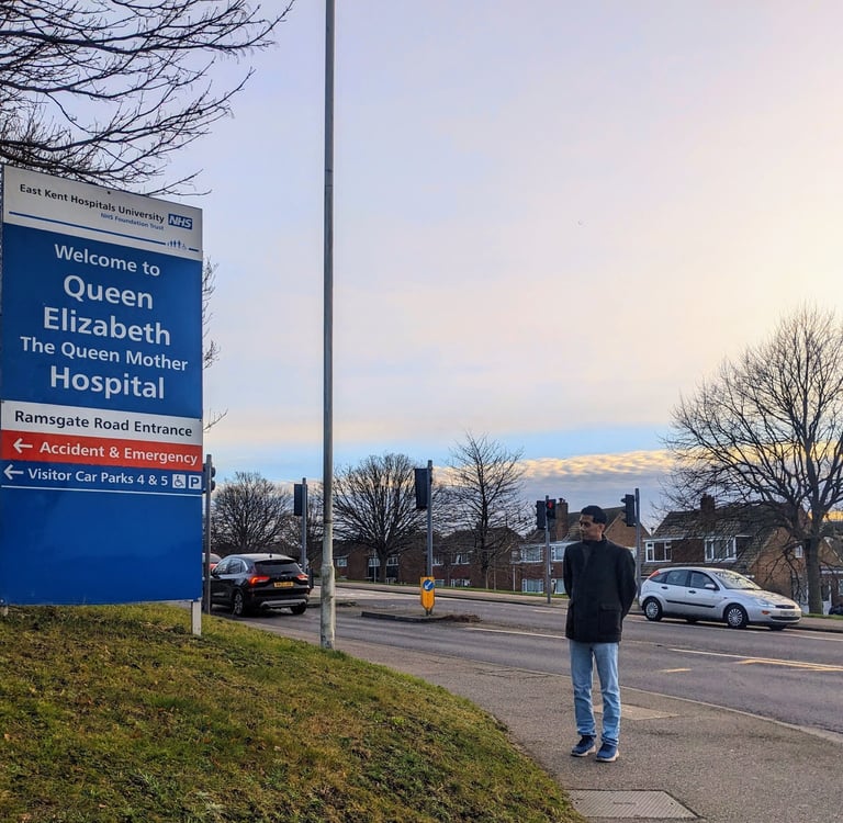 A man standing at a distance looking at the signpost reading 'Welcome to Queen Elizabeth The Queen Mother Hospital'