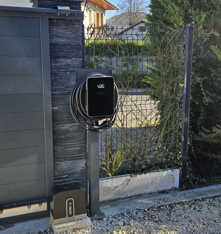 Black V2C electric vehicle wallbox charger mounted on a stone gate post in a residential driveway.