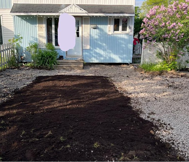 Fresh dark garden soil spread for landscaping in a gravel front yard of a blue house with pink flowers.