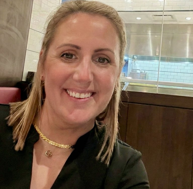 Smiling woman with blonde hair wearing a gold necklace in a modern restaurant setting.