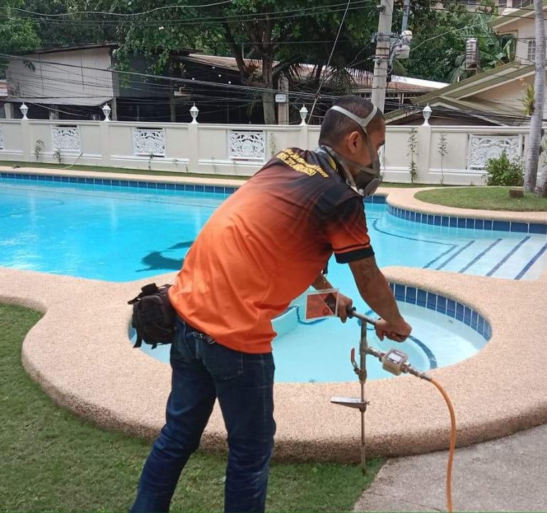 A pest control professional wearing a mask applies termite treatment near a residential swimming pool.