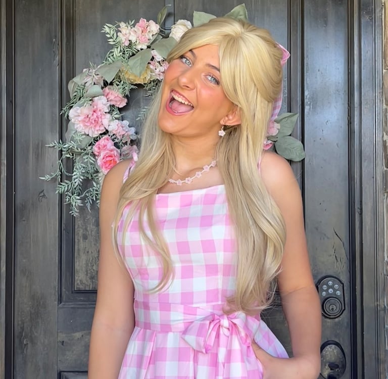 A smiling woman wearing a blonde wig and pink gingham dress in front of a front door floral wreath.