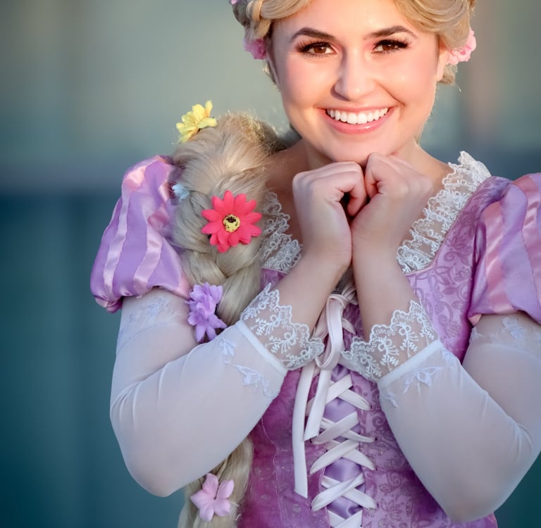 Smiling Rapunzel cosplayer wearing a purple floral dress with long braided blonde hair and flowers.