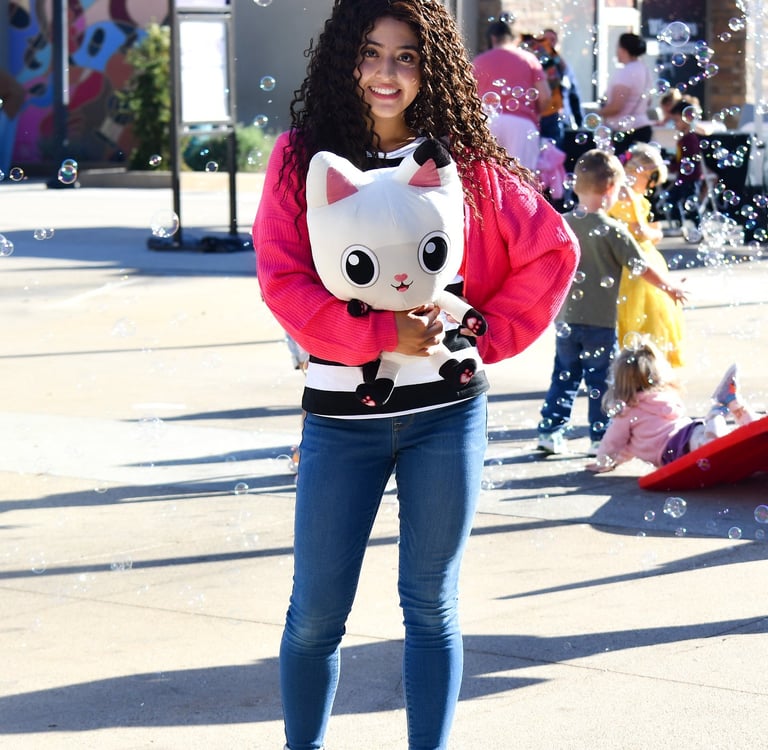 A girl in a pink sweater holds a Gabby's Dollhouse Pandy Paws plush amid floating bubbles at Jordan Landing.