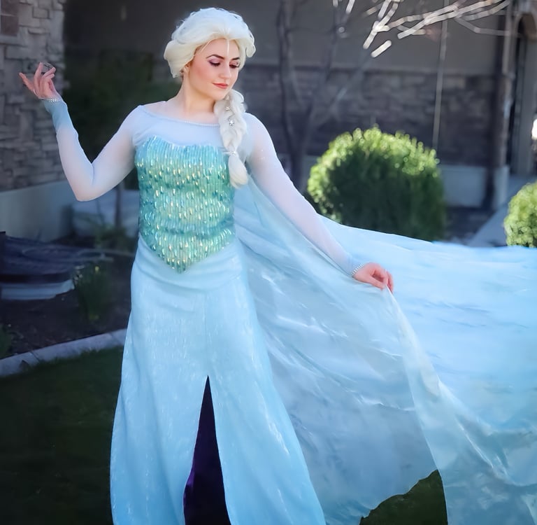 A woman in a sparkling Elsa Frozen cosplay costume with a blonde braid posing outdoors.