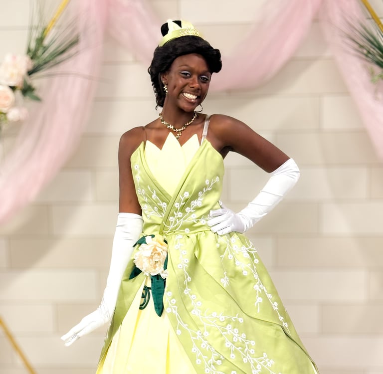 Smiling woman in a green Princess Tiana cosplay dress and white gloves posing against a floral backdrop.
